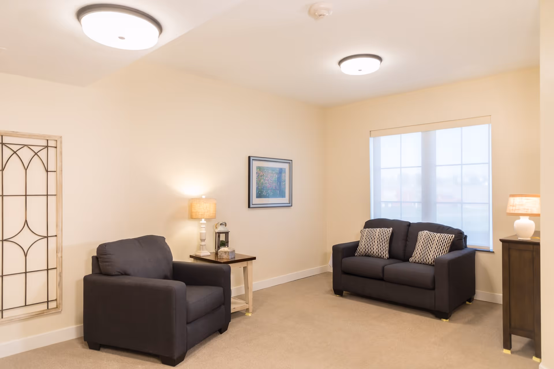 A cozy living room with beige walls and carpet, featuring a dark gray armchair and a matching two-seater sofa with patterned cushions. There are two table lamps on wooden side tables, a framed picture on the wall, and a large window with a white blind letting in natural light.