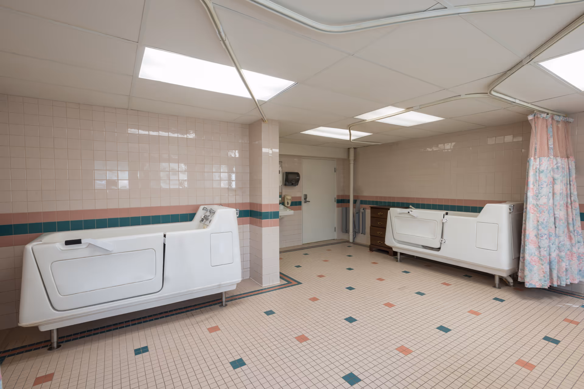 Large tiled bathing room with two white walk-in tubs, a floral privacy curtain, and pink-green floor and wall tiles.