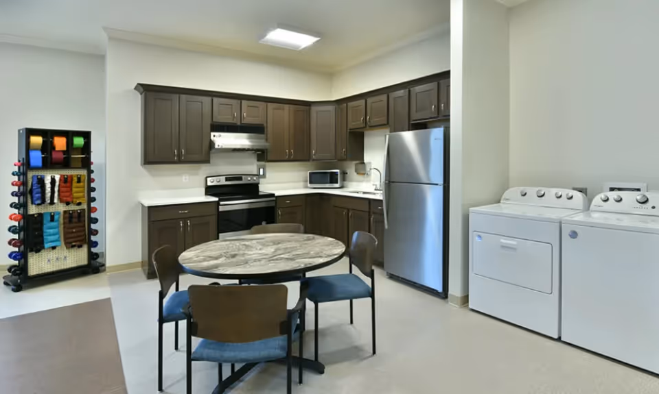 A kitchen area with dark brown cabinets, a stainless steel refrigerator, stove, microwave, and a round table with four chairs. To the right, there is a white washing machine and dryer. On the left side, there is a rack holding various colored exercise bands and weights.