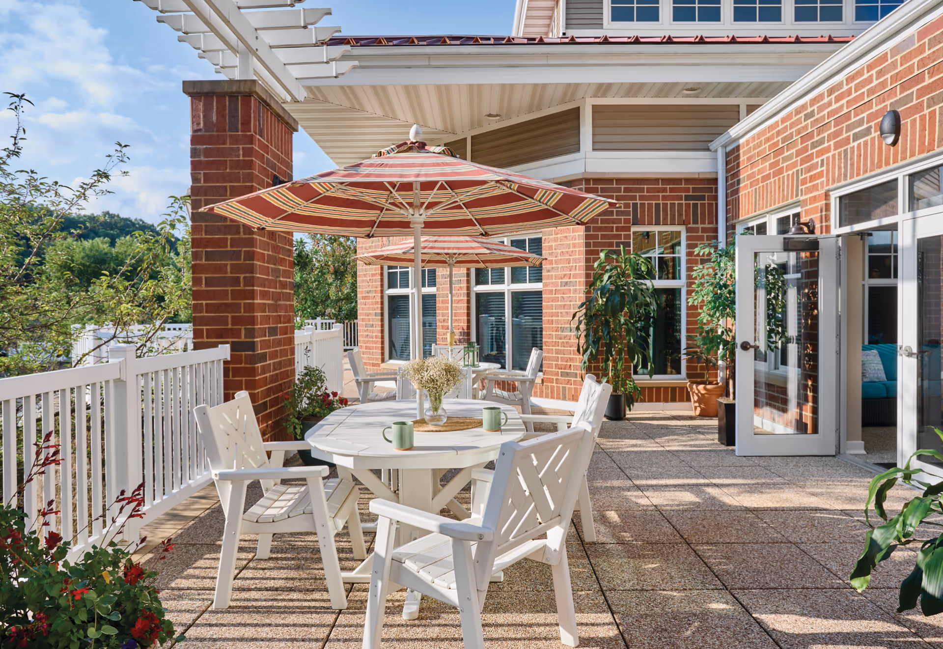 Outdoor patio area at Wittenberg Village featuring white tables and chairs with striped umbrellas, surrounded by brick walls and large windows. There are potted plants and greenery along the white railing, with a clear blue sky overhead.