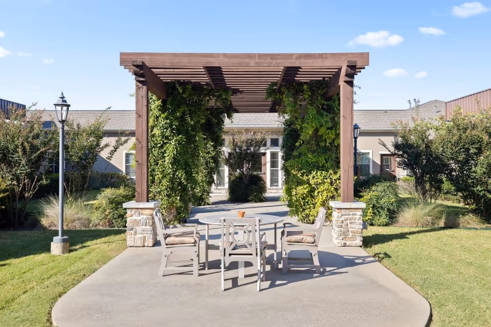 Outdoor courtyard with a wooden pergola covered in greenery and a table with chairs on a concrete patio in front of a single-story building.