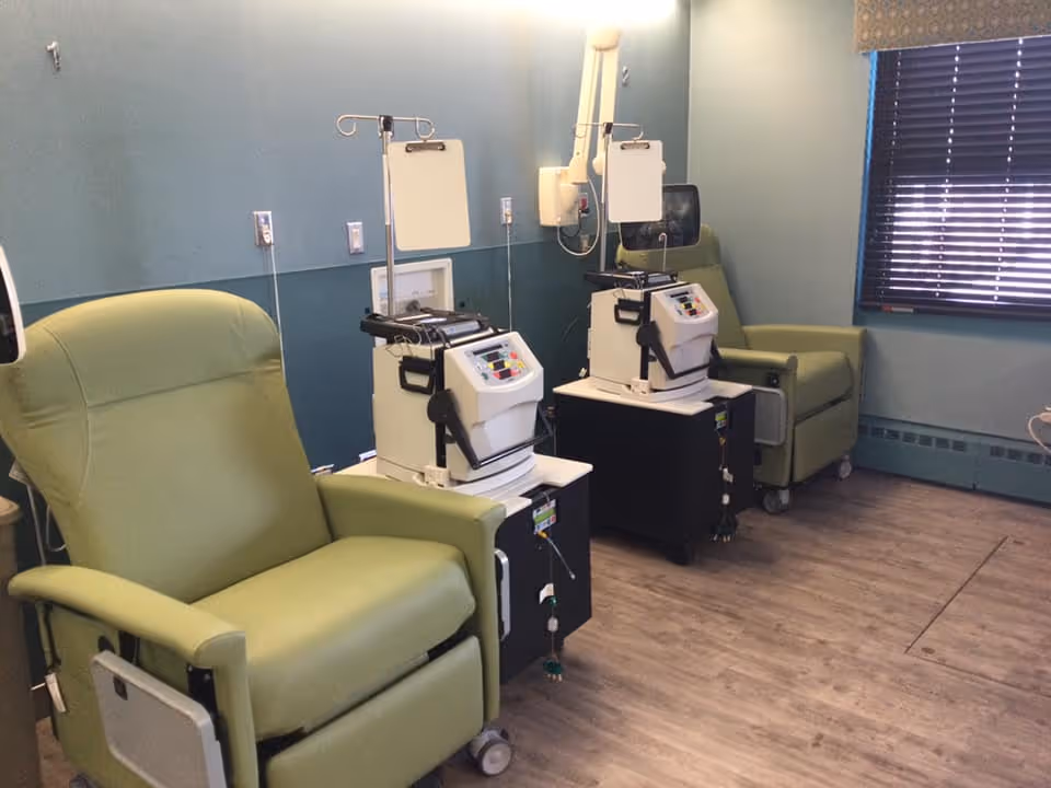 A medical treatment room with two green recliner chairs, each accompanied by medical equipment on carts with IV poles. The room has blue walls, a window with closed blinds, and wood-look flooring.