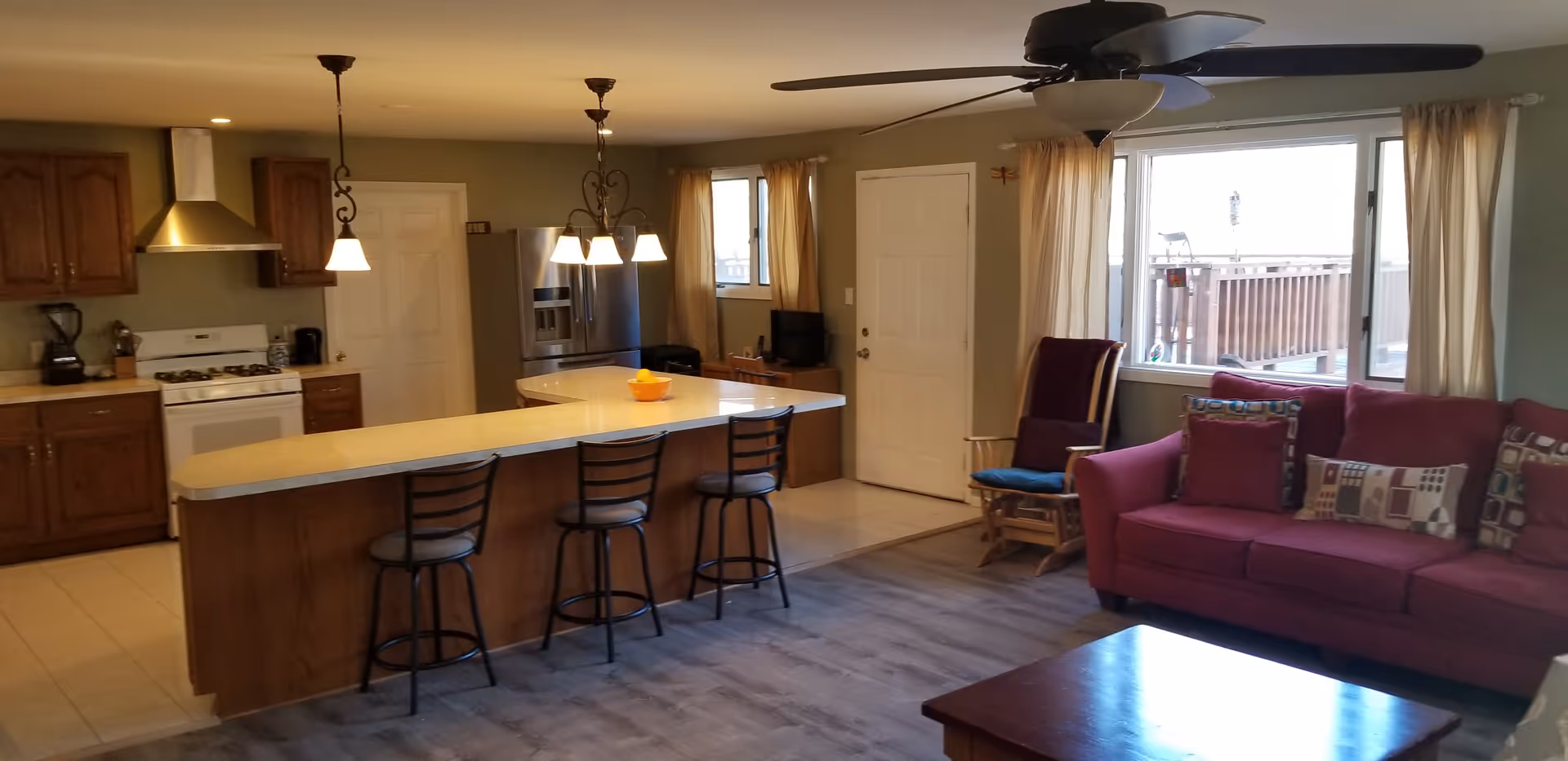 Open-concept living area with a kitchen and sitting space. The kitchen features wooden cabinets, a white stove with a stainless steel hood, a refrigerator, and a large island with three bar stools. The living area has a red sofa with patterned cushions, a wooden rocking chair with a blue cushion, a ceiling fan, and a large window with beige curtains overlooking an outdoor deck.