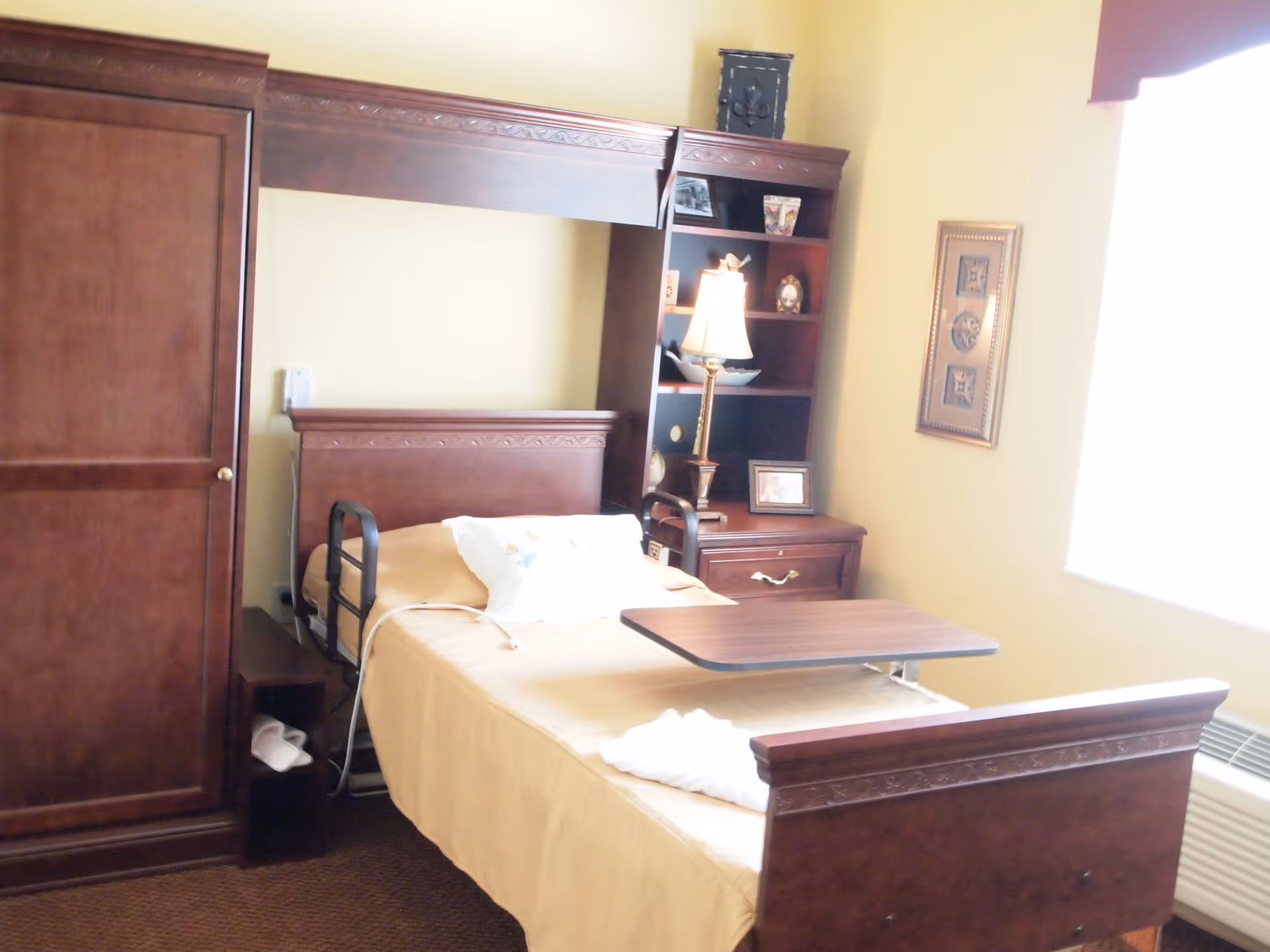A furnished bedroom with a hospital-style bed, wooden headboard and footboard, overbed table, and a bedside cabinet with lamp and shelving.