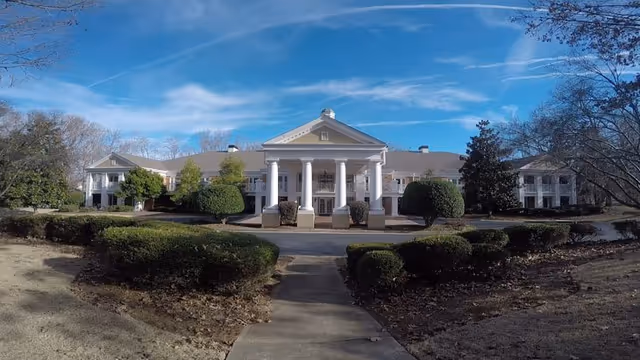 Front exterior view of a large, white, two-story building with classical architecture featuring tall white columns at the entrance, surrounded by trimmed bushes and trees under a blue sky with some clouds.