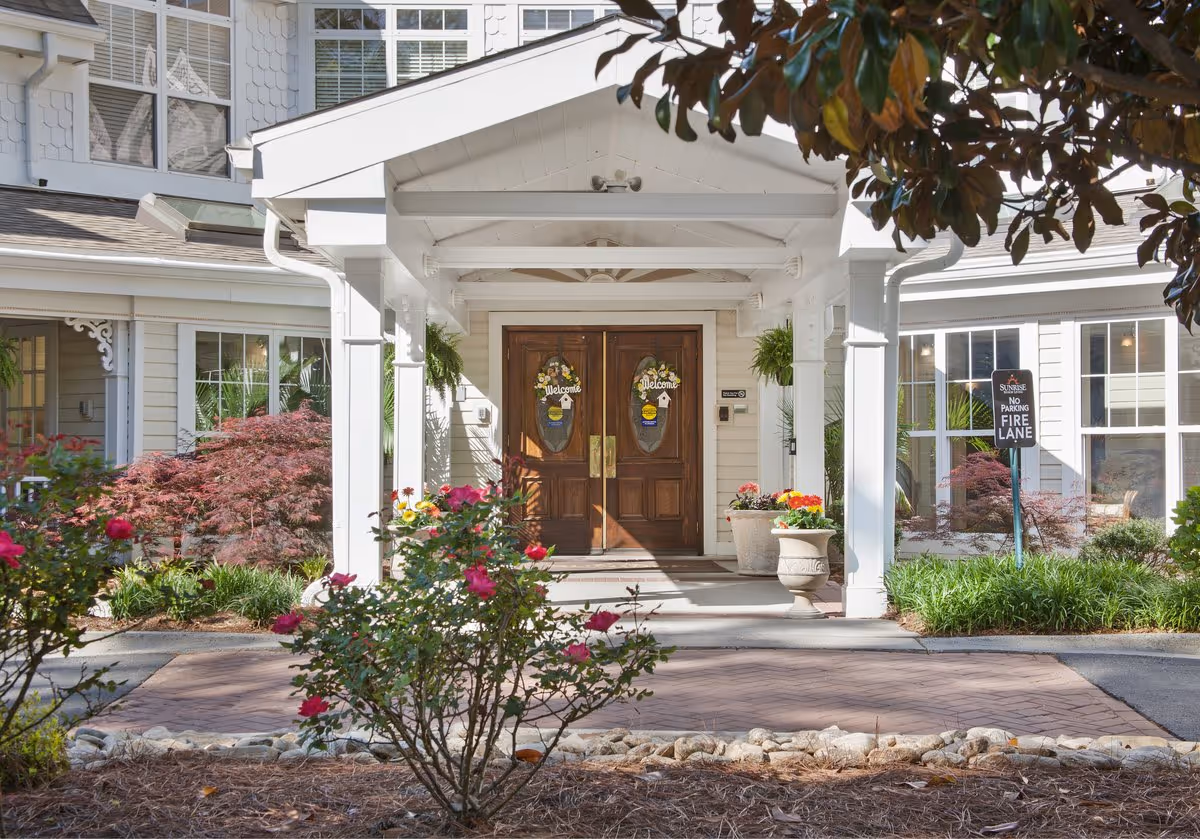Front entrance of Sunrise on Providence facility with double wooden doors decorated with welcome signs, surrounded by white pillars and a covered porch. There are colorful flowers and green plants in pots and garden beds around the entrance, with a 'No Parking Fire Lane' sign visible on the right side.