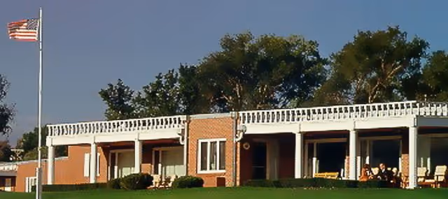 Exterior view of a single-story brick building with a white railing along the roof edge, several windows and doors, and a green lawn in front. An American flag on a flagpole is visible on the left side, and trees are in the background under a clear blue sky.