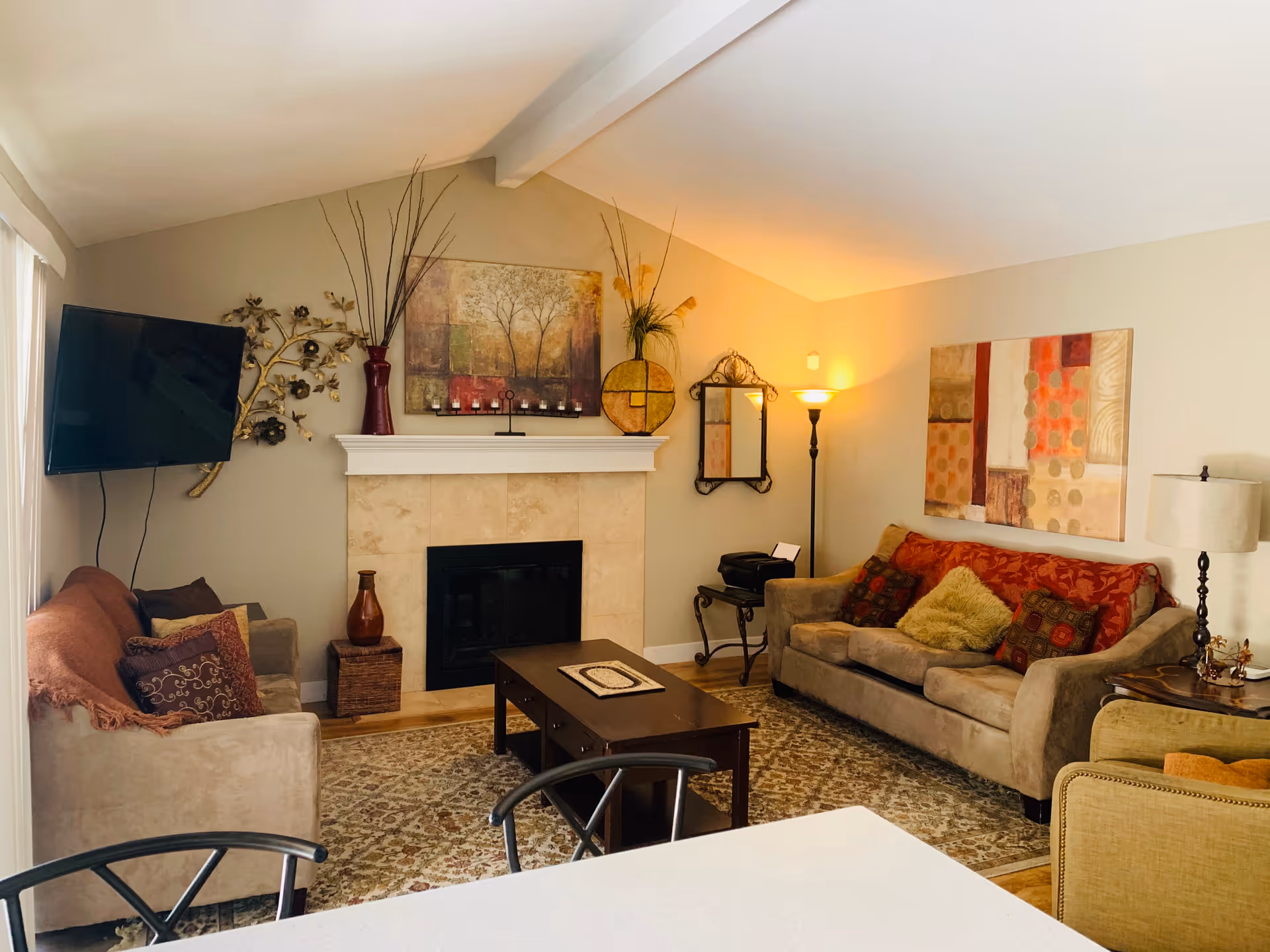 A cozy living room with beige walls and a vaulted ceiling. The room features two beige sofas with colorful cushions, a patterned area rug, a dark wooden coffee table, and a fireplace with decorative items on the mantel. A flat-screen TV is mounted on the wall to the left, and there are two standing lamps providing warm lighting. Various wall decorations and a mirror add to the room's inviting atmosphere.