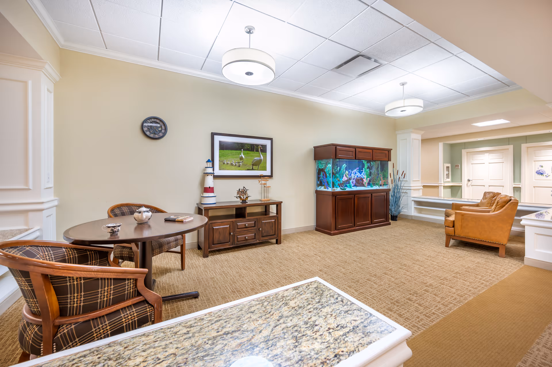 A cozy common area in a senior living facility featuring a small round table with two plaid upholstered chairs, a wooden cabinet with decorative items including a lighthouse model and a framed picture of geese on the wall above it, a large aquarium with colorful fish, and two brown leather armchairs near a hallway with white doors.
