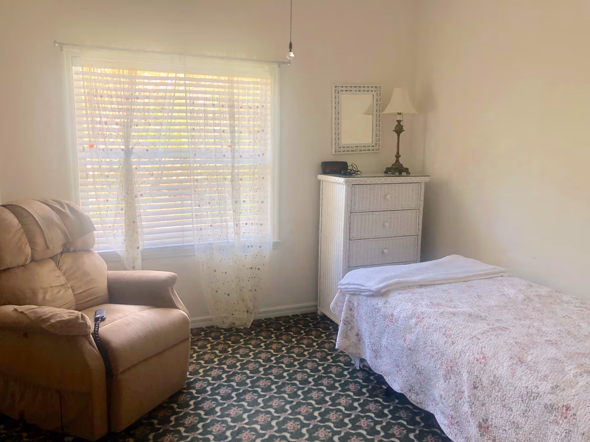 Small bedroom with a single bed, a recliner chair, a white dresser with lamp, and a window with blinds and sheer curtains.