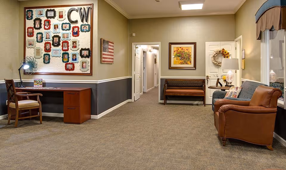 Well-lit senior living common area with a desk and chair, a wall display of framed photos, seating including a leather armchair and bench, and a hallway leading to rooms.
