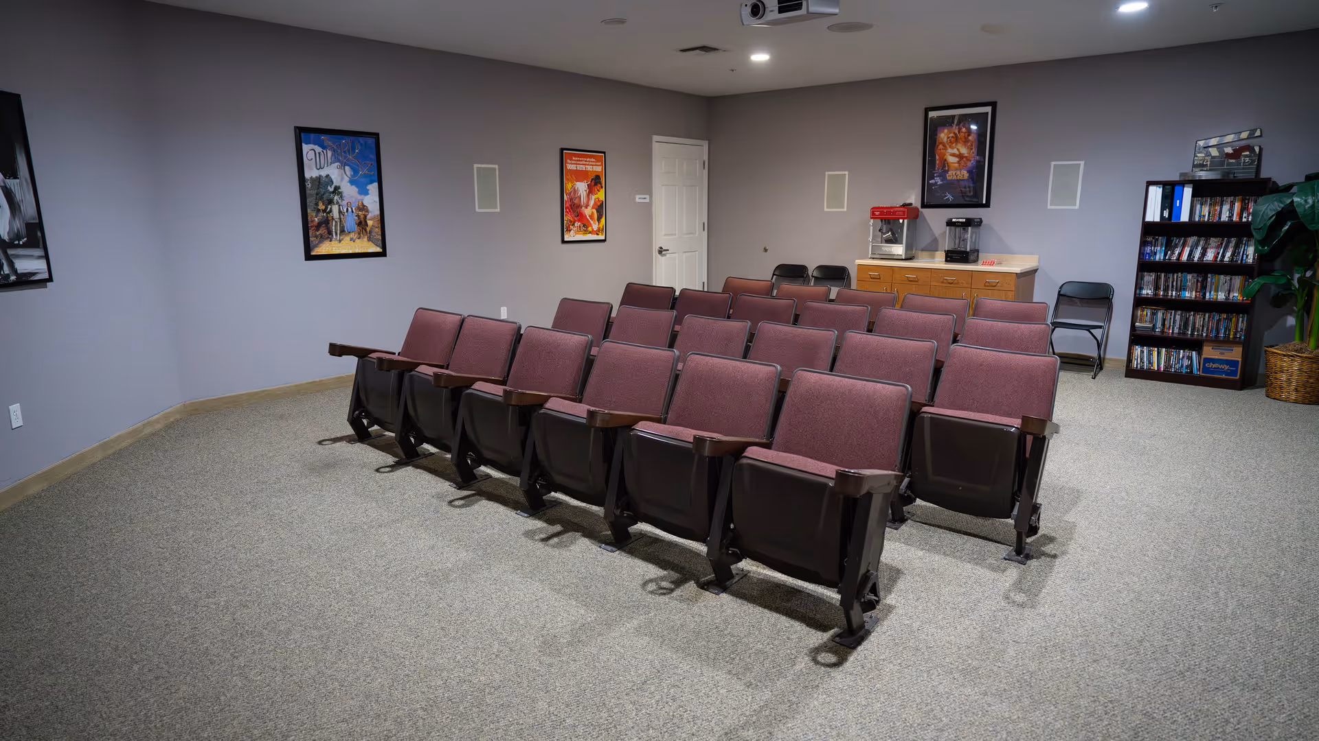 A small movie theater room with three rows of purple theater seats facing a screen. The walls are decorated with framed movie posters including The Wizard of Oz and Star Wars. There is a popcorn machine and a beverage dispenser on a counter at the back, along with a bookshelf filled with DVDs and a potted plant.
