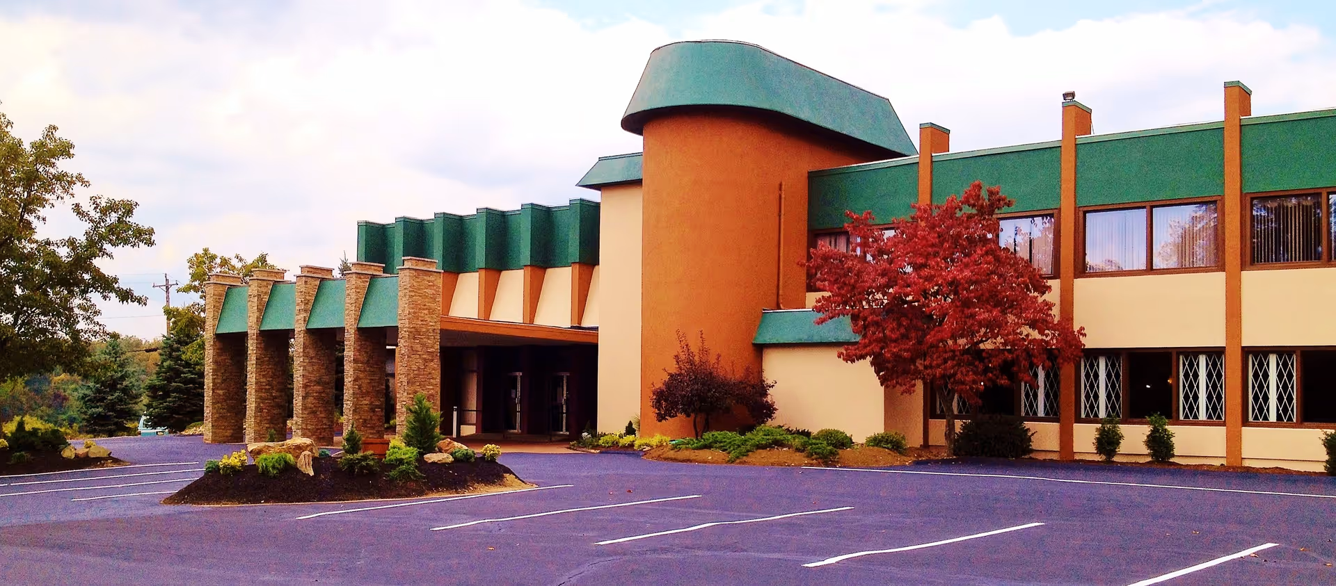 Exterior view of The Gardens of Lincoln Court building with a unique architectural design featuring stone pillars, green roof accents, and a red-leaved tree in front. The parking lot is empty and landscaped with small bushes and rocks.