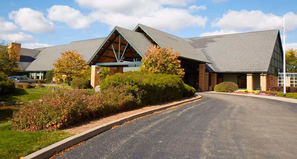 Front entrance of The Summit at Brighton Independent Living showing a covered porte-cochere, landscaped shrubs, and a curved driveway.