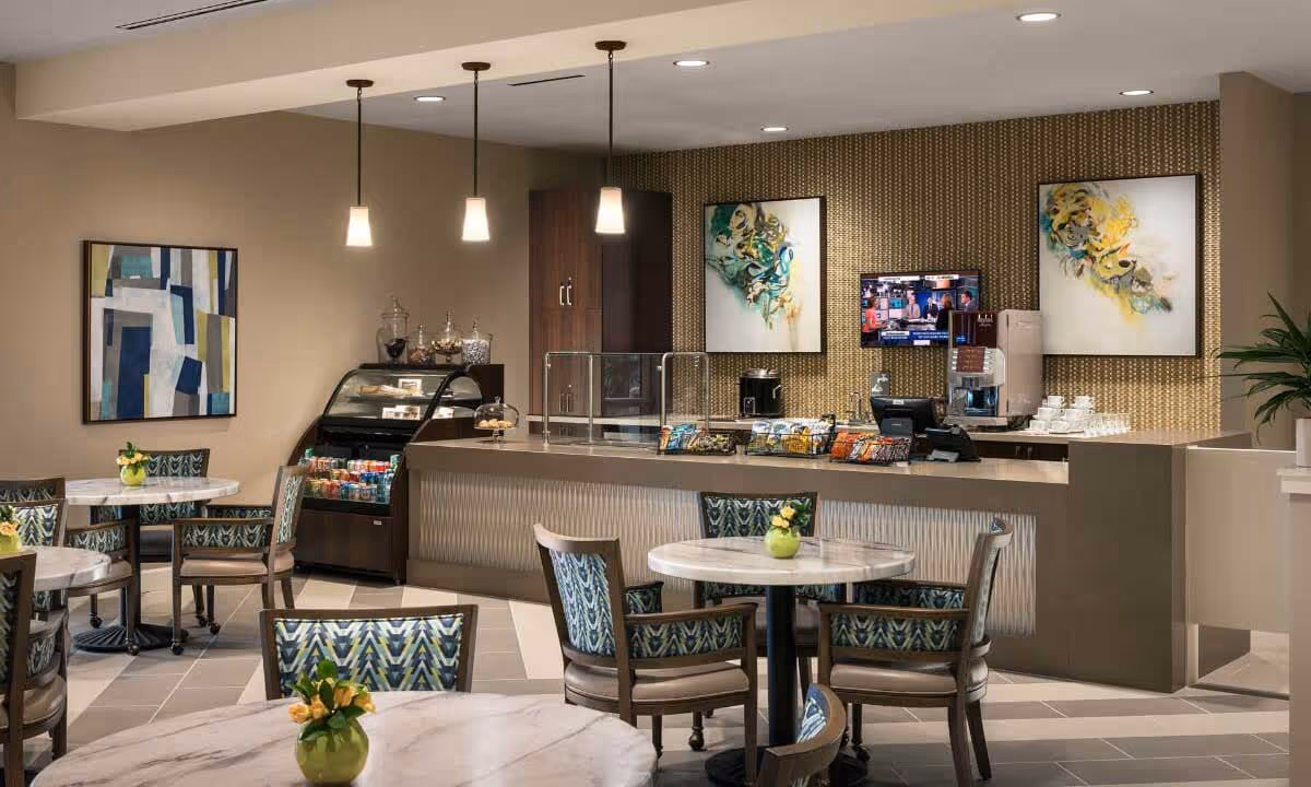 A cozy dining area in a senior living facility with round tables and patterned chairs. The room features a counter with a display case containing snacks and beverages, a coffee machine, and a small TV mounted on a textured wall. Modern pendant lights hang from the ceiling, and abstract artwork decorates the walls.