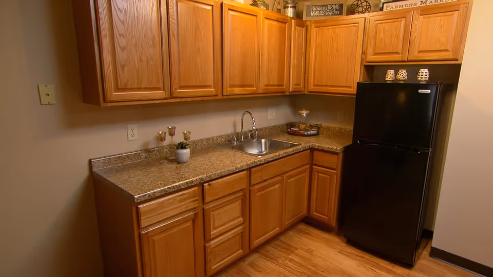 A kitchen area with wooden cabinets, a granite countertop, a stainless steel sink with a faucet, and a black refrigerator. The floor is wooden, and there are decorative items on top of the cabinets including a sign that reads 'Farmers Market'.