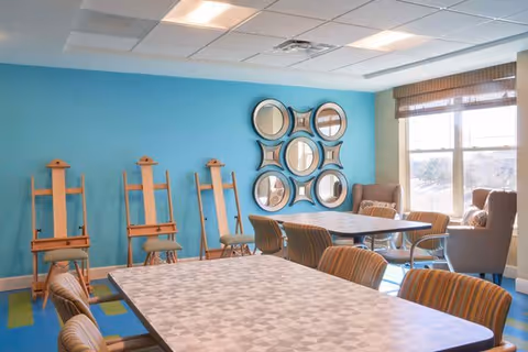 A bright room with a blue accent wall featuring six circular mirrors arranged in a square pattern. There are four wooden easels lined up against the wall and two tables with patterned surfaces surrounded by striped chairs. Two armchairs are positioned near a large window with blinds, letting in natural light.