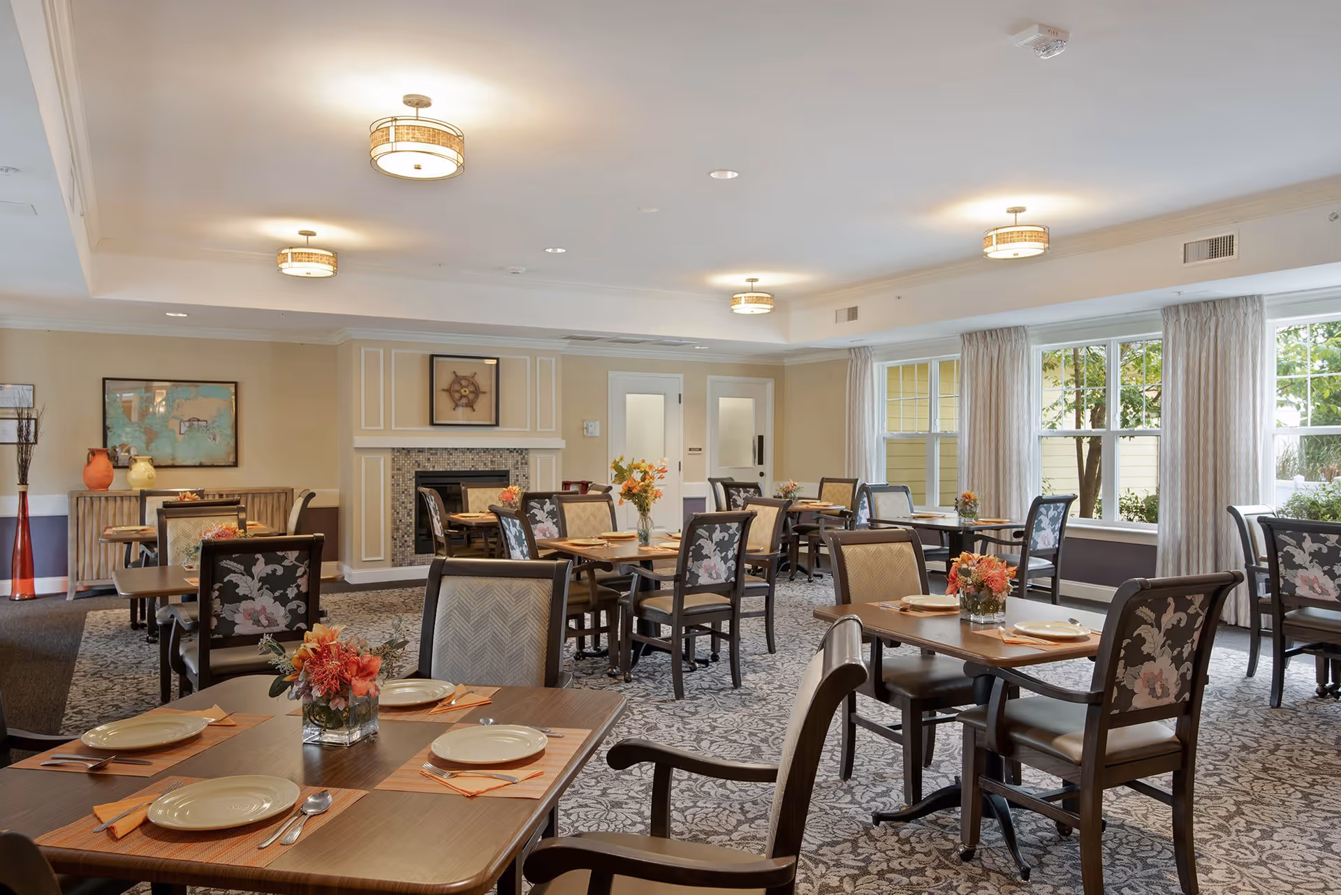 A well-lit dining room in a senior living facility with multiple wooden tables and chairs arranged neatly. Each table is set with plates, utensils, and floral centerpieces. Large windows with curtains allow natural light to brighten the room. The walls are decorated with framed artwork and a fireplace is visible in the background.