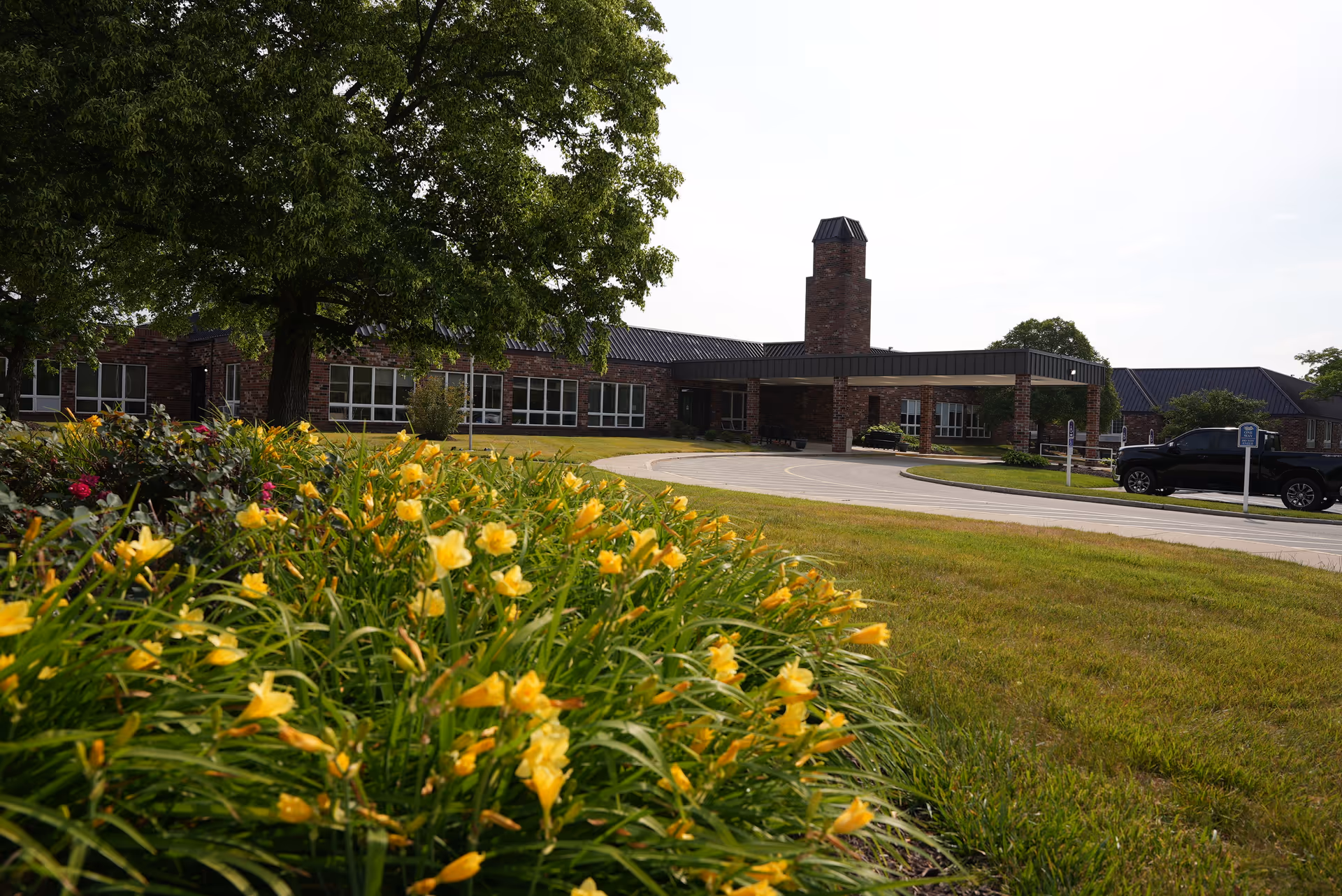 Front entrance of a brick senior living facility with a circular driveway, parked car, large tree, and yellow flowers in the foreground.
