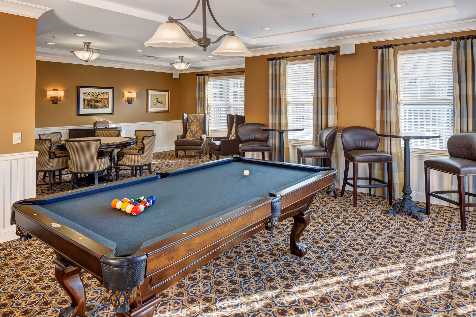 A well-lit recreational room with a pool table in the center, set up for a game with balls racked and a cue ball on the table. The room features patterned carpet, mustard-colored walls with white wainscoting, and several windows with plaid curtains allowing natural light. There are multiple seating areas including a round table with chairs, high tables with bar stools, and two upholstered armchairs near the windows. Ceiling lights and wall sconces provide additional lighting.