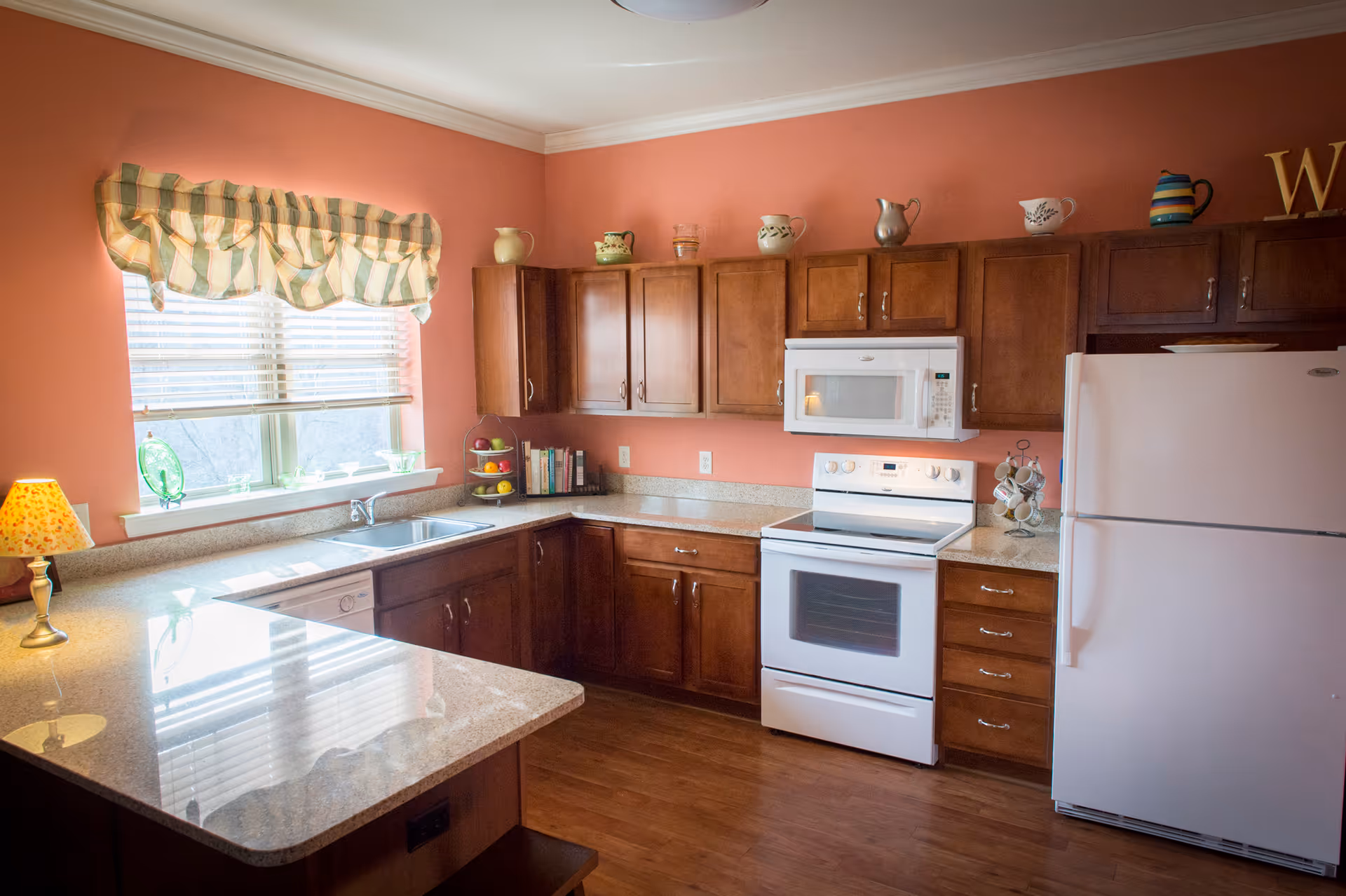 A cozy kitchen with wooden cabinets, a white refrigerator, white stove with an overhead microwave, and a double sink under a window with striped valance curtains. The walls are painted a warm coral color and the countertops are light-colored granite. Decorative pitchers and mugs are displayed on top of the cabinets, and a small lamp with a patterned shade sits on the counter.