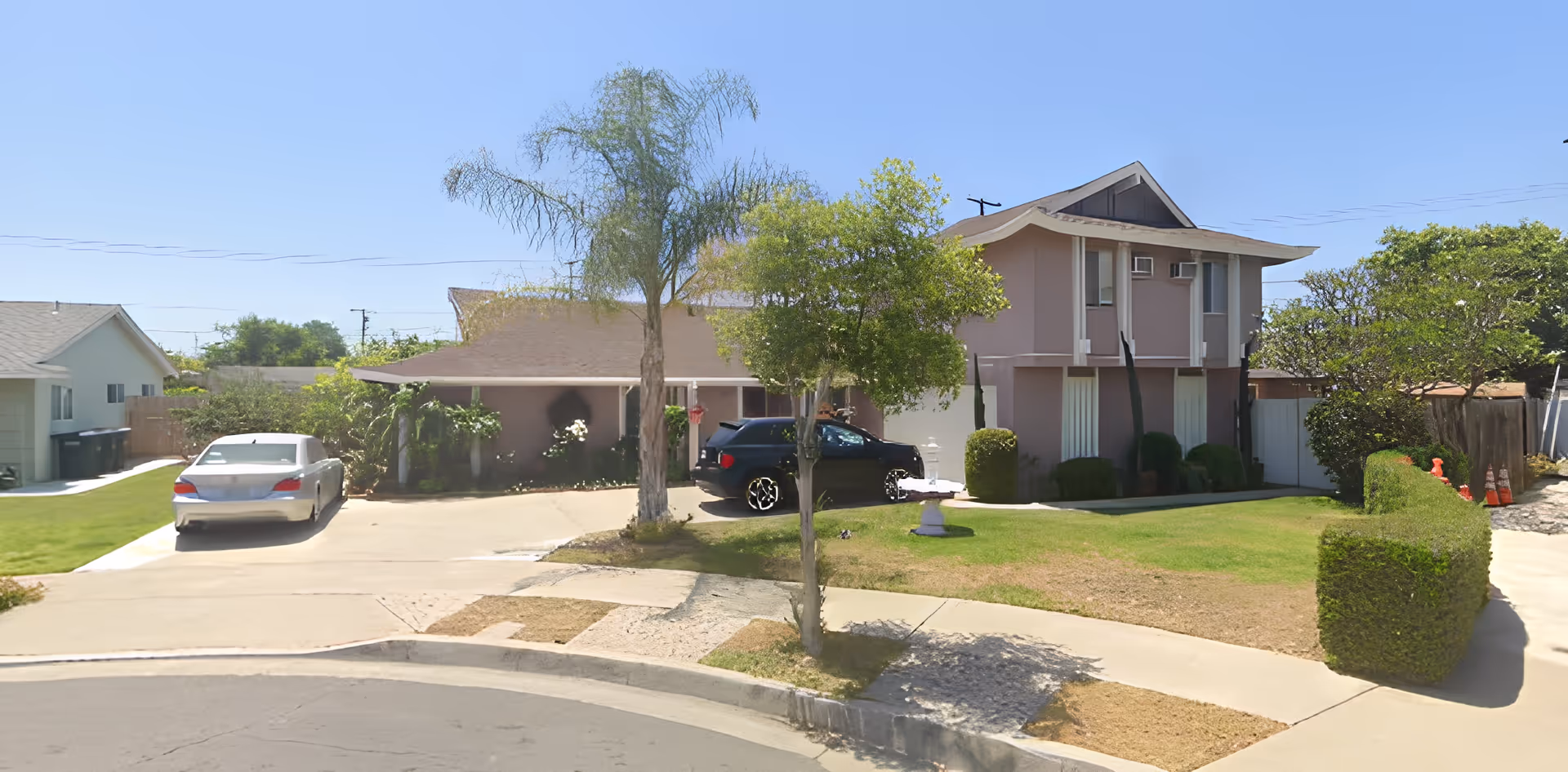 Exterior view of a two-story residential building with a driveway, two parked cars, green lawn, trees, and bushes under a clear blue sky.