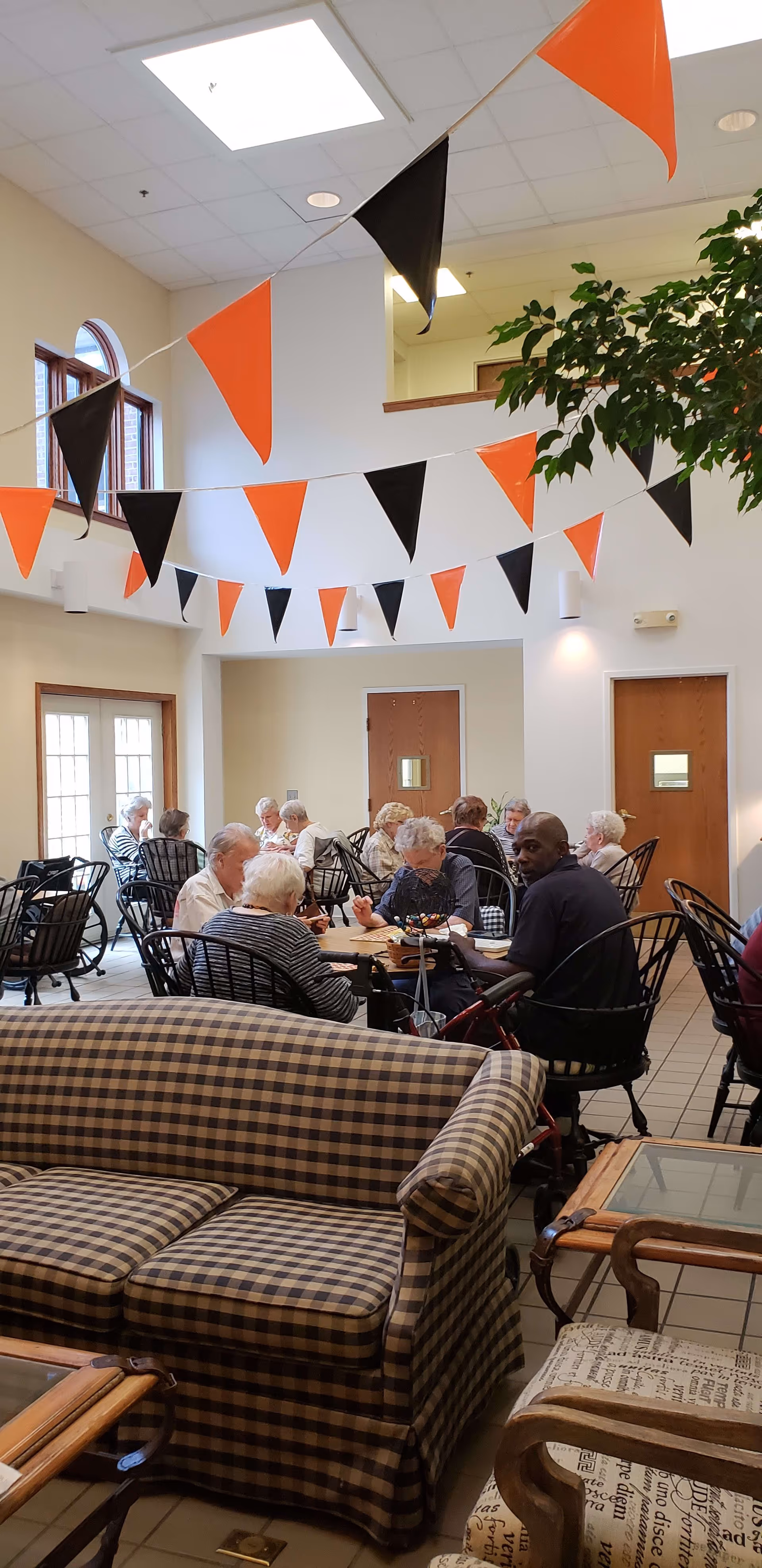 A group of elderly people seated at tables in a bright, decorated common dining area with checkered couches and orange-and-black pennant banners overhead.