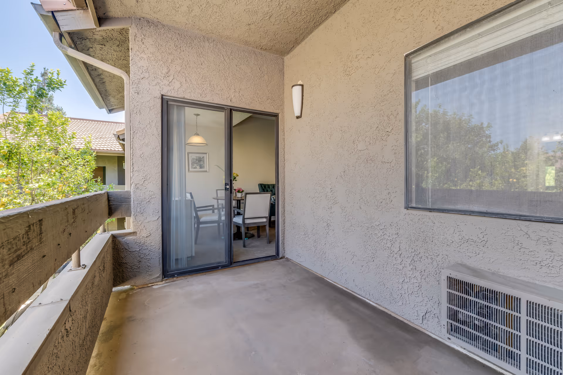A covered outdoor balcony area with textured beige walls, a sliding glass door leading to an interior dining area with a table and chairs, a wall-mounted light fixture, and a window with a view of trees outside.