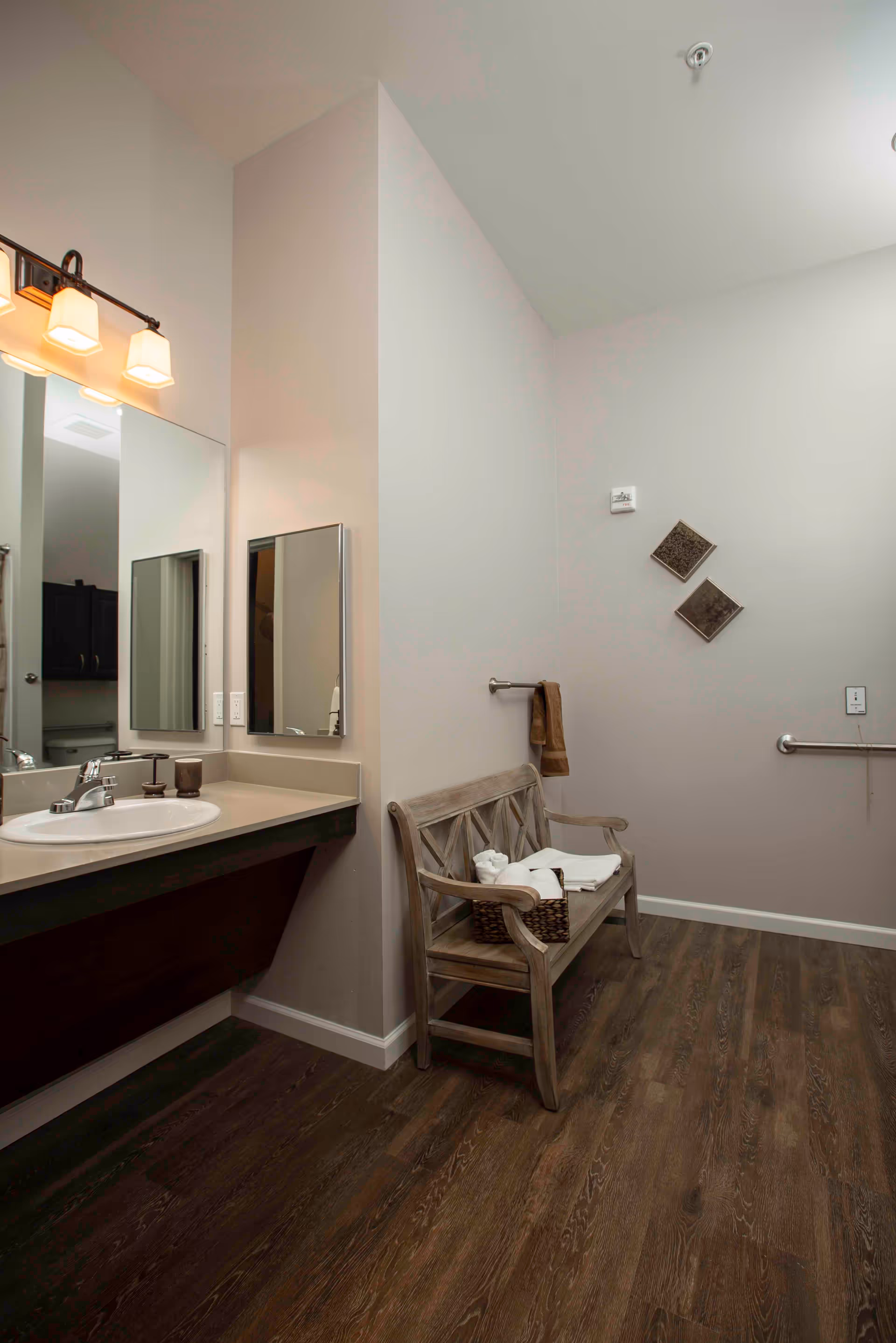 Accessible bathroom with a sink and mirror on the left, a wooden bench holding towels in the center, and grab bars on the right wall.