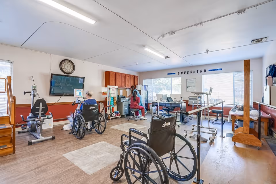 A rehabilitation room in a senior living facility with exercise equipment, wheelchairs, parallel bars for walking support, and two people engaged in therapy activities. The room has large windows letting in natural light, a wall clock, and cabinets along one wall. A motivational phrase 'SUPERHERO' is displayed on the wall.