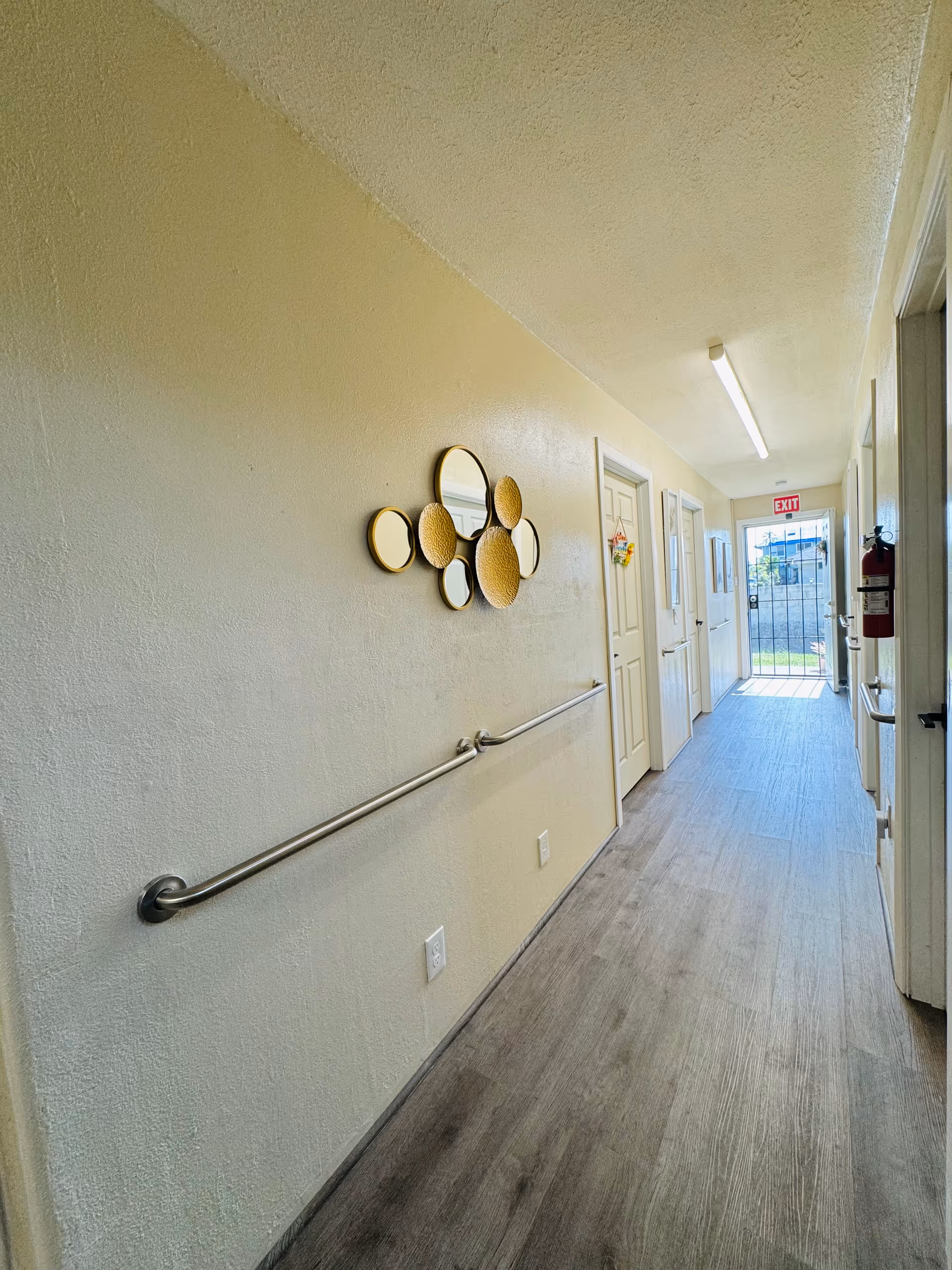 A well-lit hallway with beige walls and wood-look flooring. There are handrails on both sides of the hallway, decorative round mirrors on the left wall, several closed white doors on the right, and an exit door with a red exit sign at the far end. A fire extinguisher is mounted on the right wall near the exit.