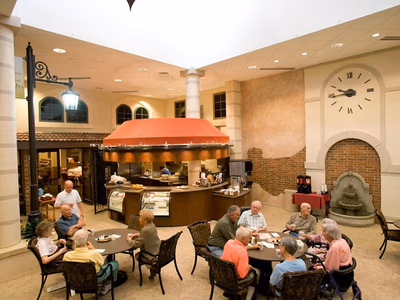 A group of elderly people sitting around two tables in a spacious indoor dining area with a high ceiling. There is a central food service counter with a red canopy, a large clock on the wall, and a decorative water fountain. The atmosphere is warm and inviting with soft lighting.