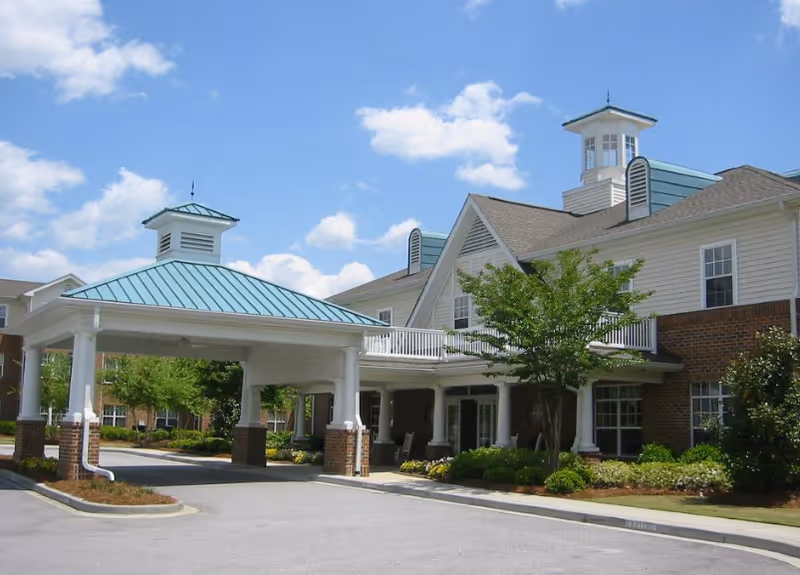 Exterior view of a senior living facility named Wildewood Downs featuring a covered entrance with white columns and a teal metal roof. The building has beige siding with brick accents, multiple windows, and a small tower structure on the roof. There are trees and shrubs around the entrance and a clear blue sky with some clouds above.