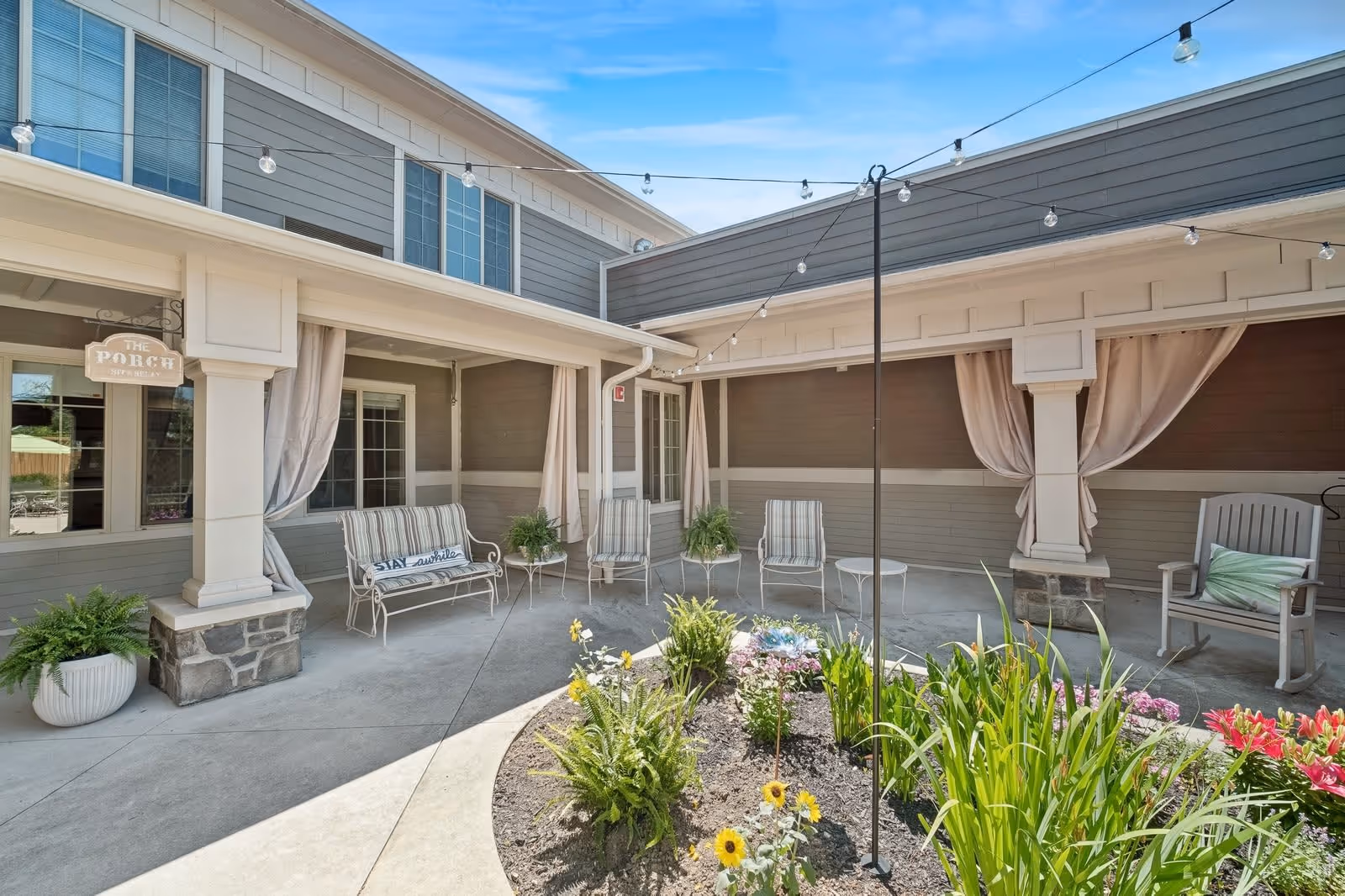 Outdoor courtyard area with a garden bed in the center featuring various flowers and plants. Surrounding the garden are several chairs and benches, some with cushions, under a covered porch with beige curtains tied back. String lights are hung above the seating area. The building exterior is gray with white trim.