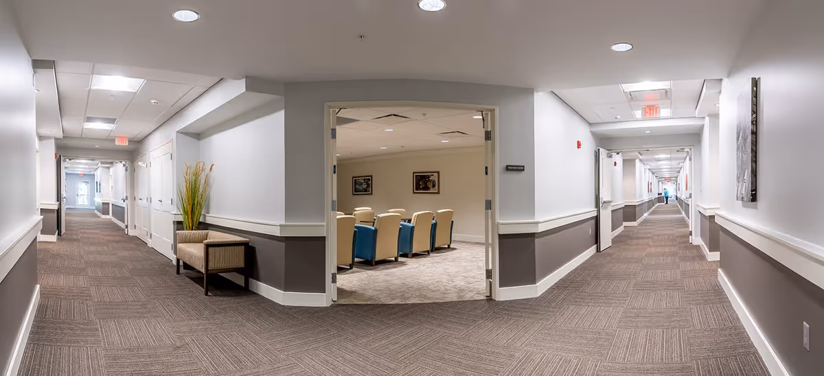 A wide hallway in a senior living facility with beige and brown carpeting and white walls with a gray lower section. The hallway has several closed doors on both sides and a small seating area with a chair and a tall plant on the left. In the center, double doors open into a room with beige and blue chairs arranged in rows. The hallway extends into the distance with ceiling lights and exit signs visible.