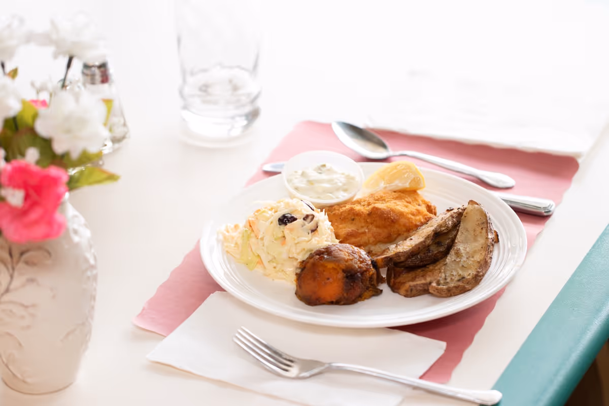 A plate with fried fish, potato wedges, coleslaw and a roll on a table set with utensils and a vase of flowers.