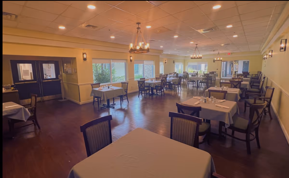 A spacious dining room in a senior living facility with multiple tables covered with white tablecloths, each set with glasses, napkins, and silverware. The room has wooden chairs, large windows letting in natural light, and warm ceiling lights including chandeliers. The floor is wooden, and the walls are painted a light color with sconces providing additional lighting.