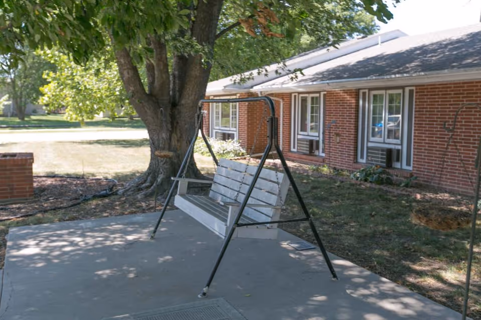 Outdoor patio area with a white wooden swing bench hanging from a black metal frame, situated on a concrete slab. A large tree provides shade, and a brick building with multiple windows is visible in the background.