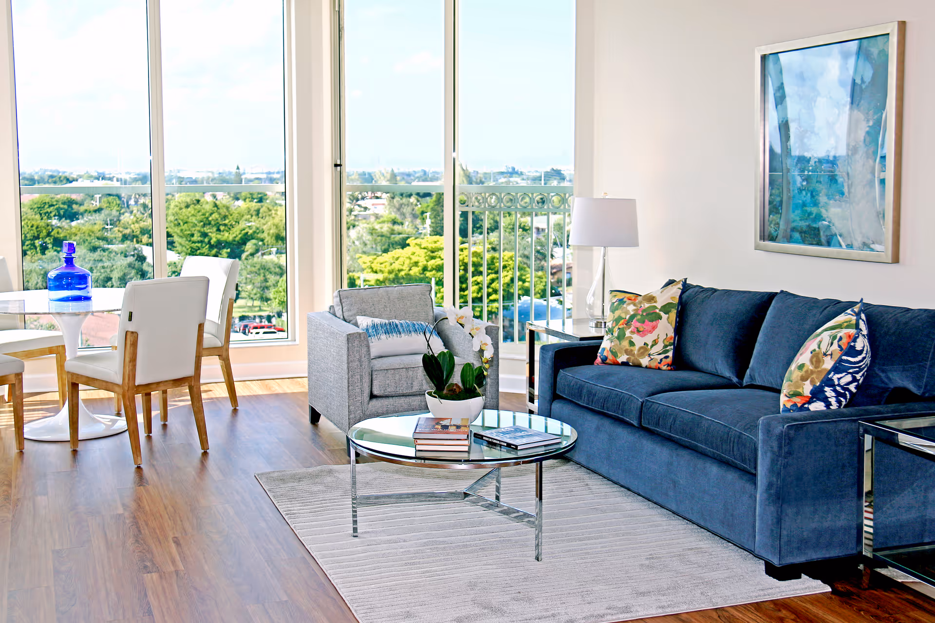 A bright living room with large windows showing a green outdoor view. The room features a blue sofa with floral and patterned cushions, a gray armchair with a blue throw, a glass coffee table with books and a potted plant, a side table with a lamp, and a round dining table with white chairs and a blue glass bottle on top.
