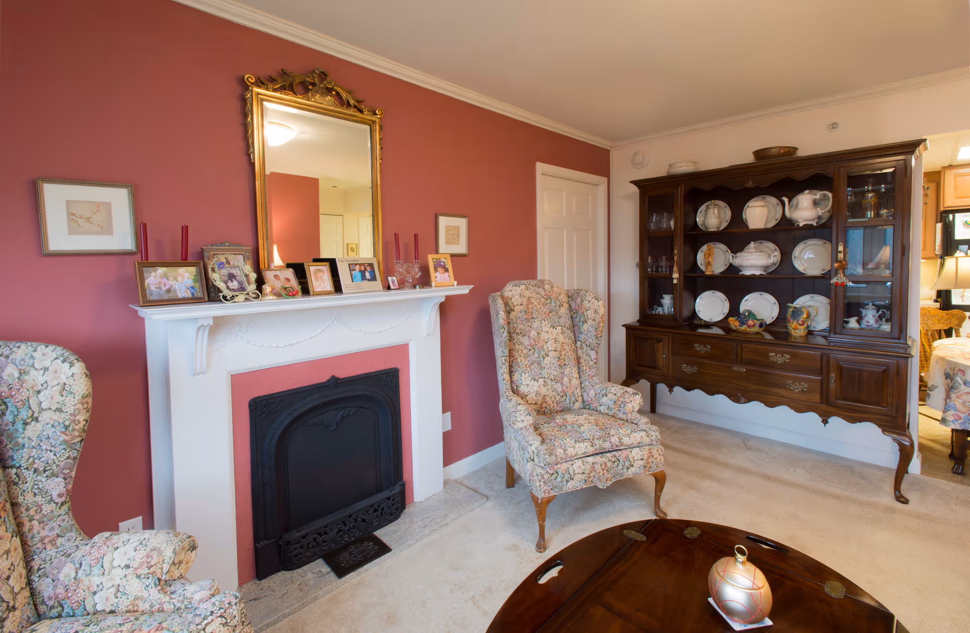 Cozy living room with floral armchairs, a white mantel fireplace topped by a gold-framed mirror, and a wooden china cabinet displaying plates.