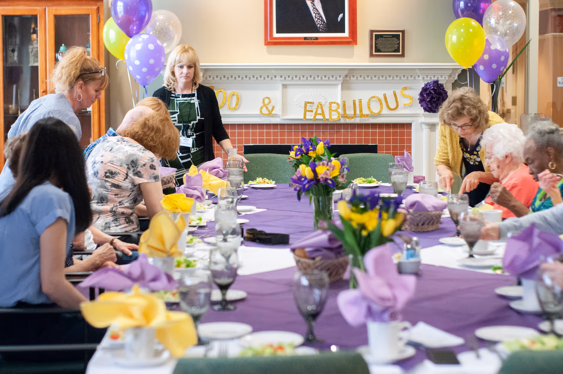A group of elderly people and caregivers gathered around a long dining table decorated with purple tablecloth, yellow and purple napkins, and flower centerpieces. Balloons and a banner reading '100 & FABULOUS' are displayed on the wall behind them, indicating a celebration.