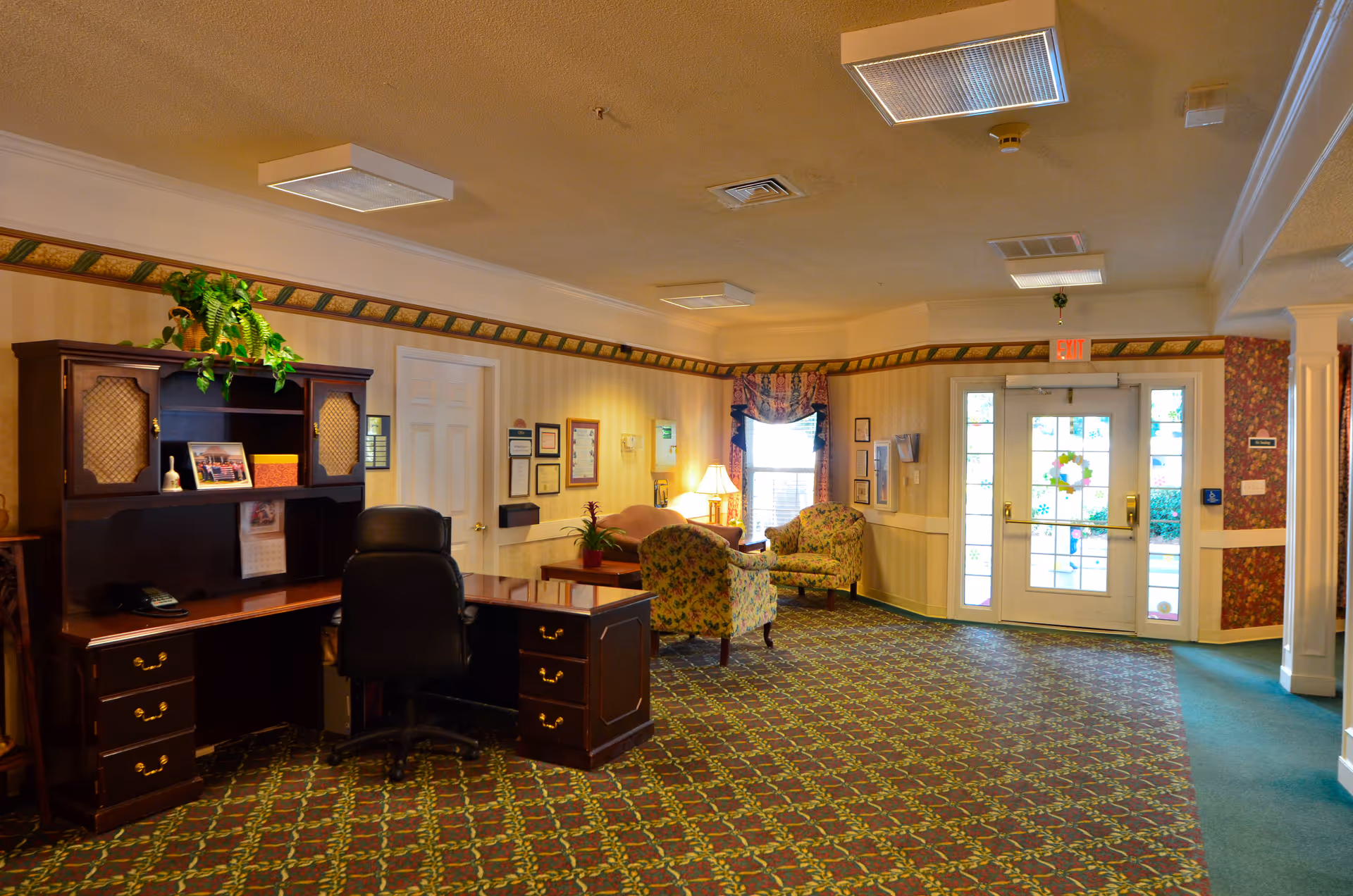 Well-lit senior living facility lobby with a reception desk, armchairs, patterned carpet, and glass double doors.