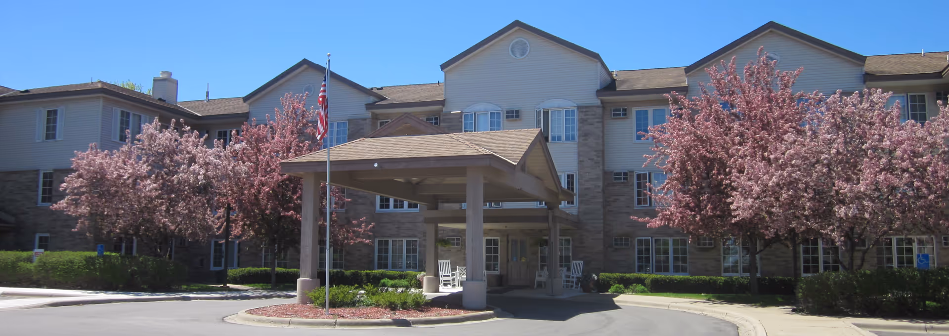Front exterior view of a senior living facility named Elim Shores with a covered entrance, American flag, blooming pink trees, and a clear blue sky.