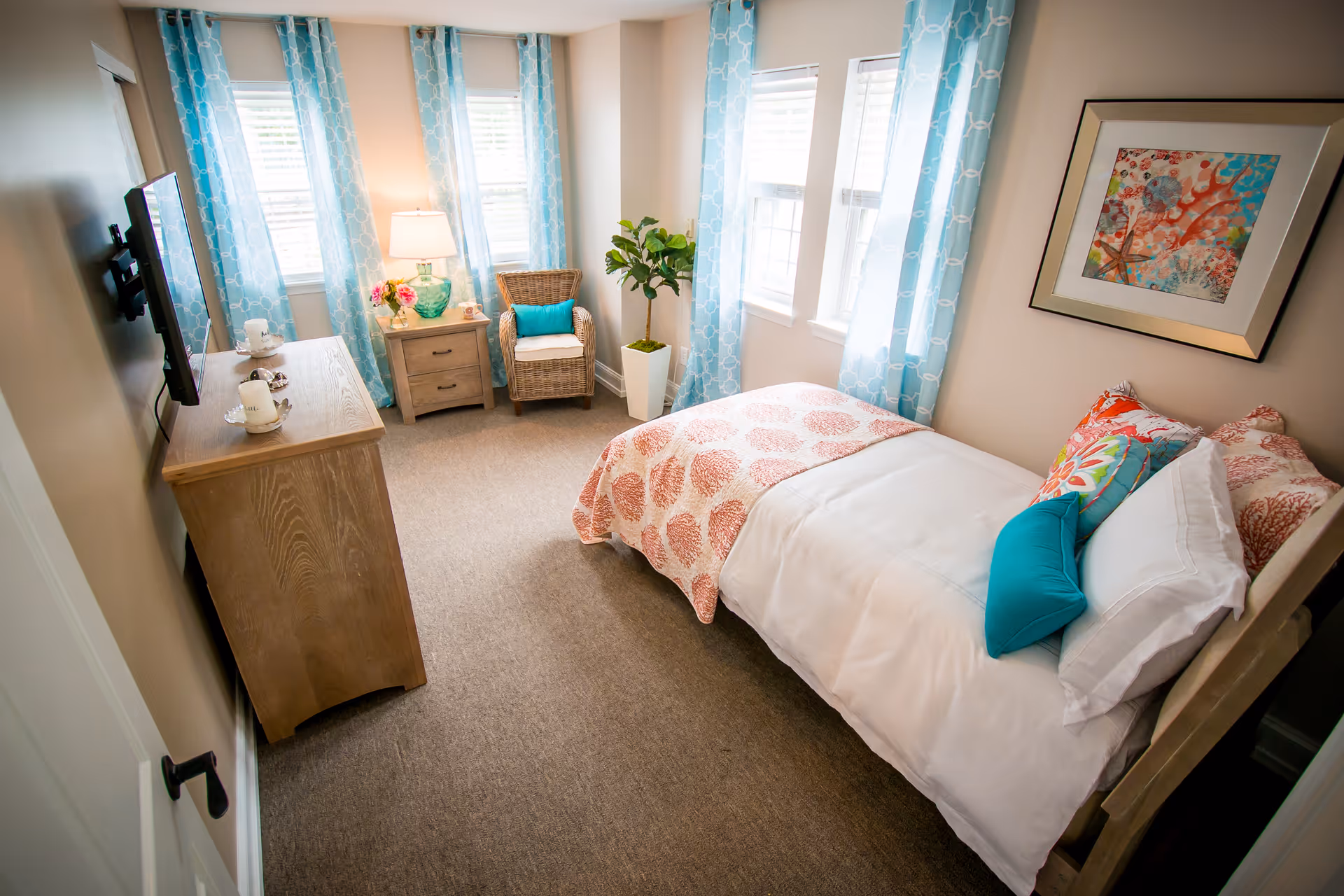 Bright furnished bedroom with a single bed, dresser and wall-mounted TV, a wicker chair by windows with blue curtains, and decorative artwork.