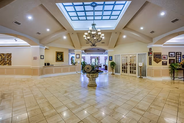 Spacious senior living lobby with a central ornamental fountain, tiled floor, chandelier under a skylight, and a reception desk area.