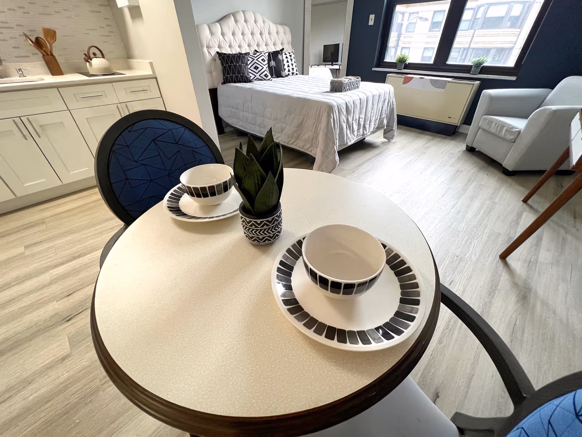 Studio apartment interior showing a small round dining table set for two, a bed, kitchenette cabinets, and a chair by a window.