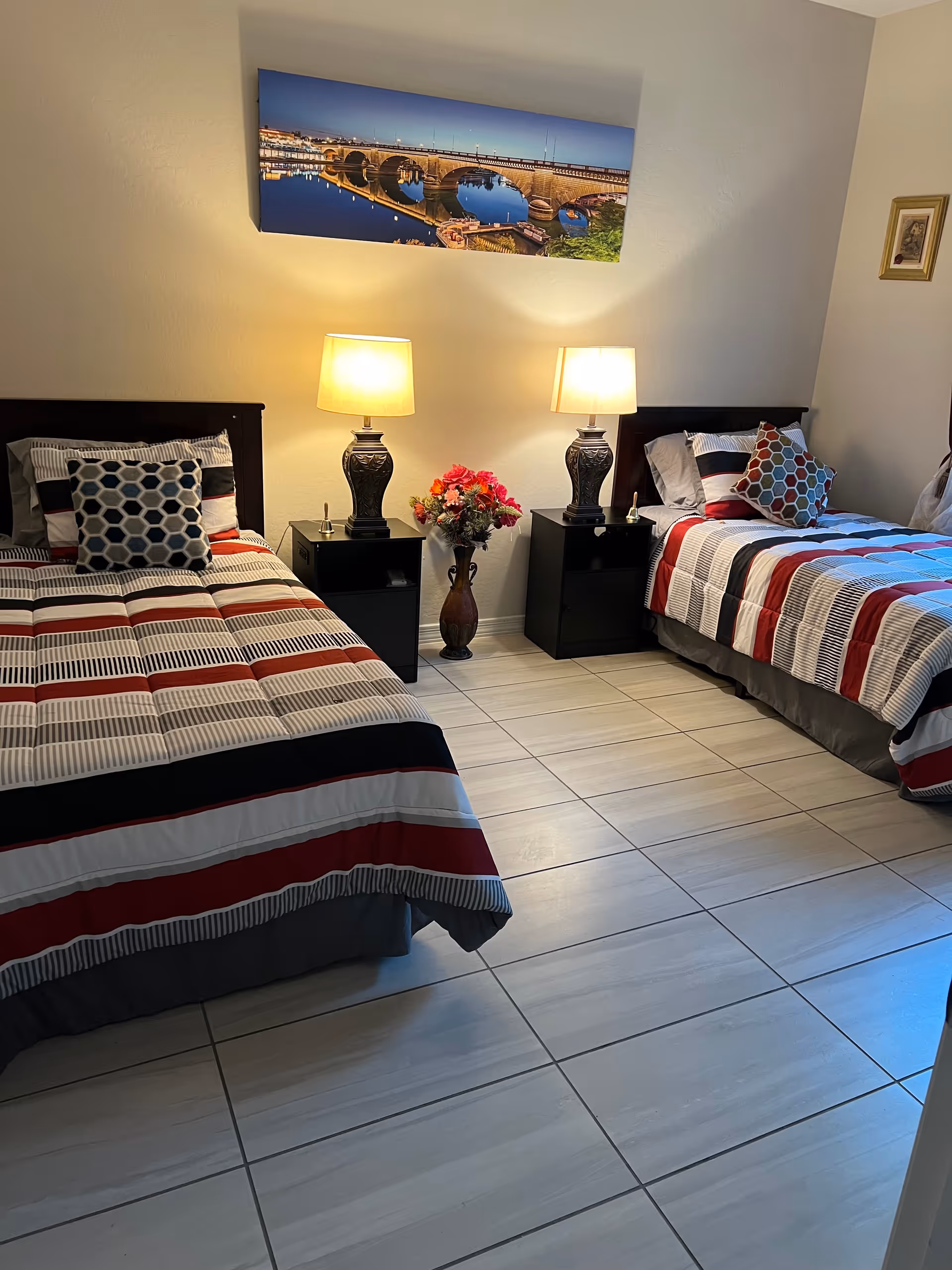 A bedroom with two single beds, each with striped bedding in red, white, black, and gray colors. Each bed has a decorative pillow with a hexagonal pattern. Between the beds are two black nightstands, each with a lamp featuring a dark base and a light-colored shade. A vase with colorful flowers is placed on the floor between the nightstands. Above the beds, a large framed picture of a bridge over water at dusk is hung on the wall. The floor is tiled with light-colored tiles.