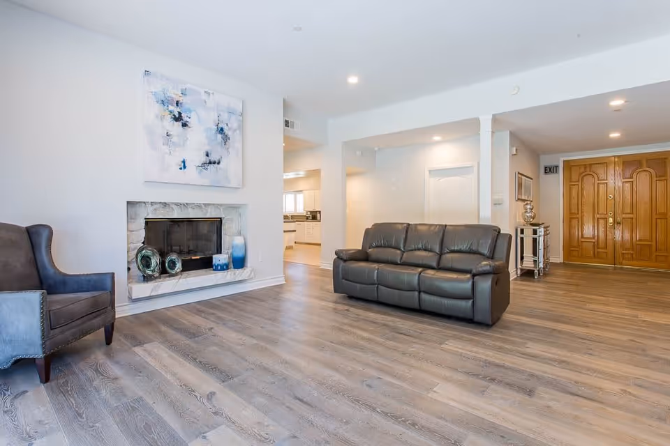 A spacious living room with light wood flooring, a dark leather sofa, a gray armchair, and a fireplace with decorative items on the mantel. There is a large abstract painting above the fireplace, a small mirrored side table with a silver lamp, and double wooden doors in the background.