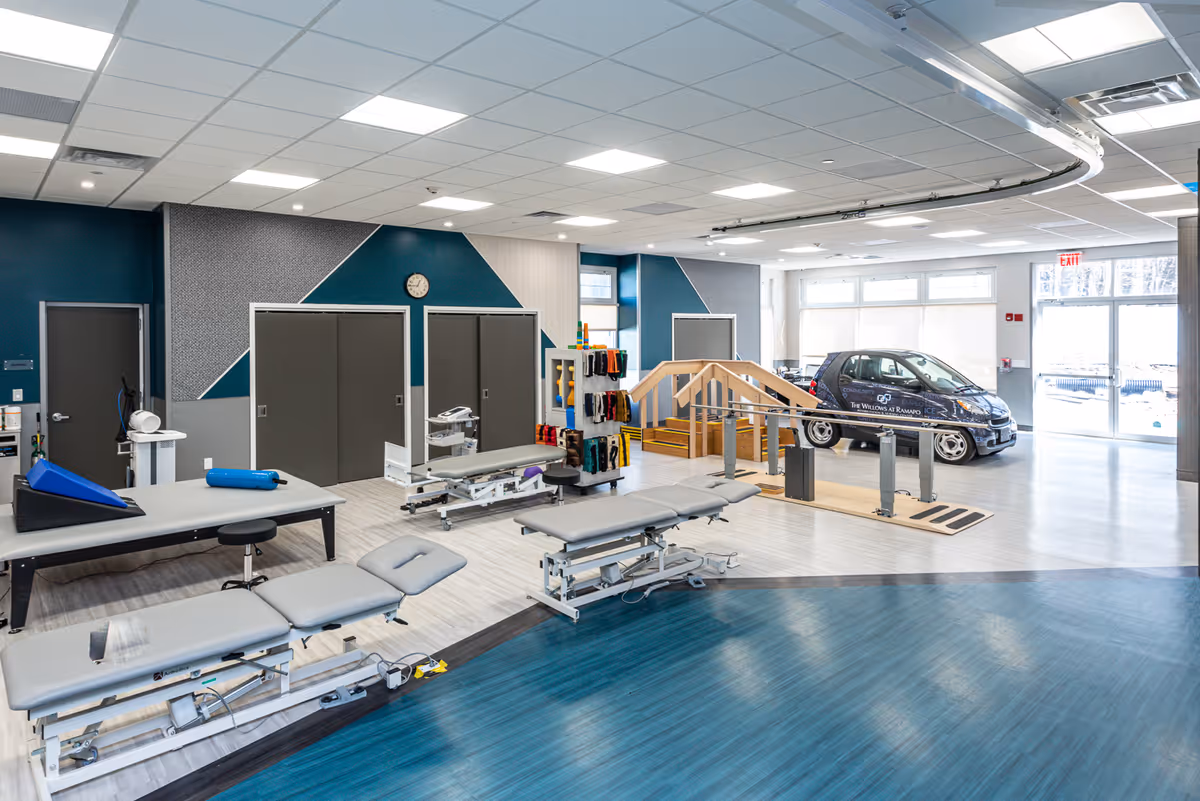 A spacious rehabilitation therapy room with multiple adjustable treatment tables, therapy equipment, and a small car used for mobility training. The room has large windows allowing natural light, a clock on the wall, and a wooden ramp with handrails for physical therapy exercises.