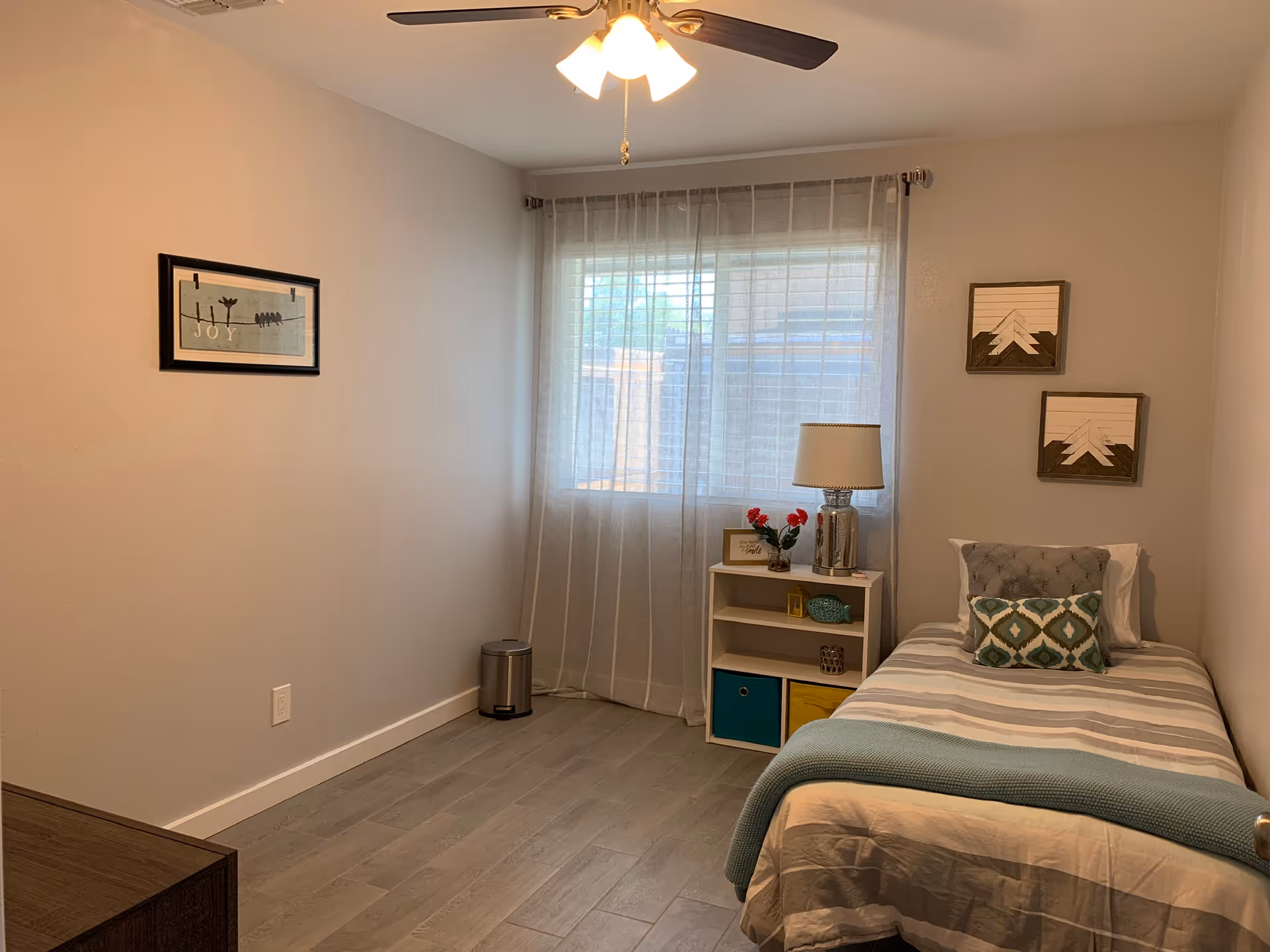 A small, neatly arranged bedroom with a single bed against the right wall. The bed has a striped bedspread, a blue throw blanket, and two decorative pillows. Next to the bed is a small white shelving unit with a lamp, flowers, and decorative items. A window with sheer curtains allows natural light into the room. The walls are light-colored and decorated with three framed artworks. A ceiling fan with lights is visible above.
